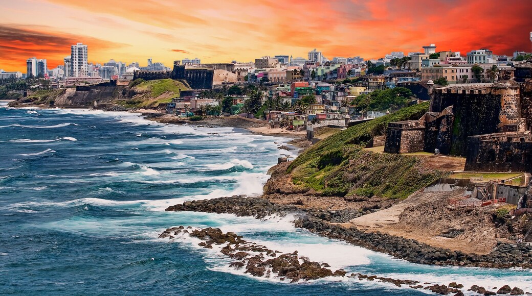A coastal cityscape with waves crashing against rocky shores, historic buildings, and a sunset sky.