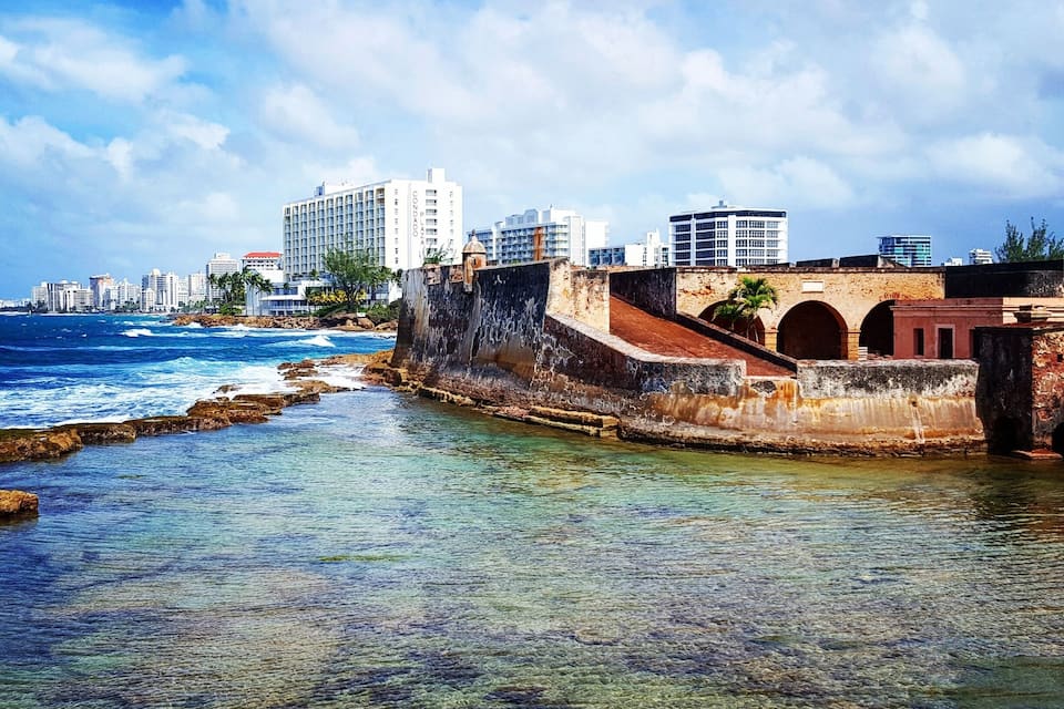 Un paisaje urbano costero con aguas azul cristalino, un muro de piedra histórico y edificios modernos altos.