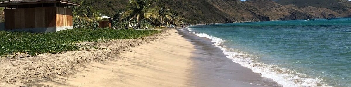 A sandy beach with a wooden structure, palm trees, and mountains in the background.