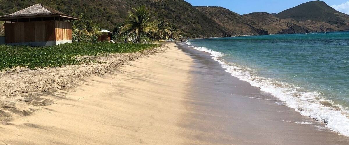 Une plage de sable avec une structure en bois, des palmiers et des montagnes en arrière-plan.