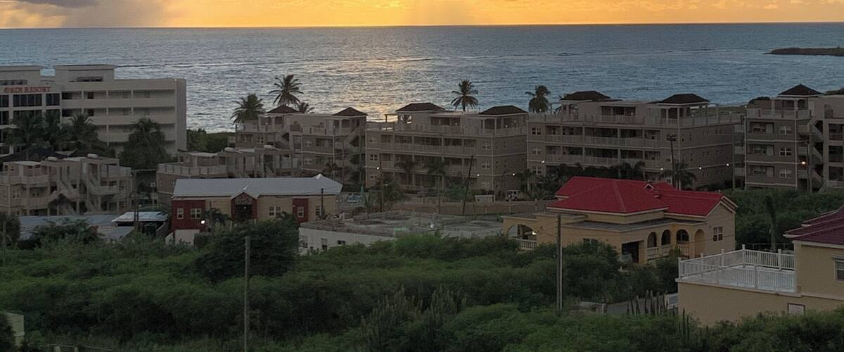 A coastal cityscape with buildings, palm trees, and a sunset over the ocean.