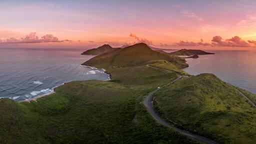 A coastal landscape with lush green hills, a winding road, and a sunset over the ocean.