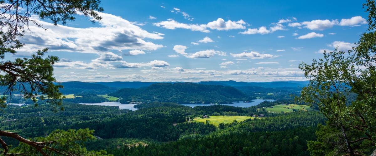 A mountainous landscape with dense forests, a lake, and distant hills under a partly cloudy sky.