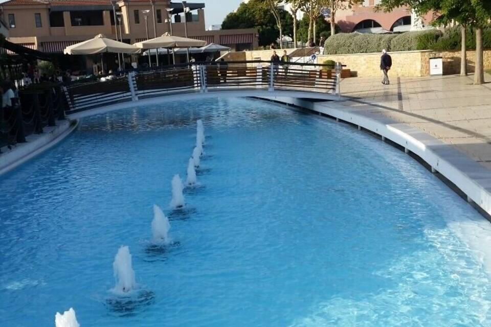 Uma piscina com vários jatos de água, rodeada por um espaço pavimentado com árvores e edifícios ao fundo.