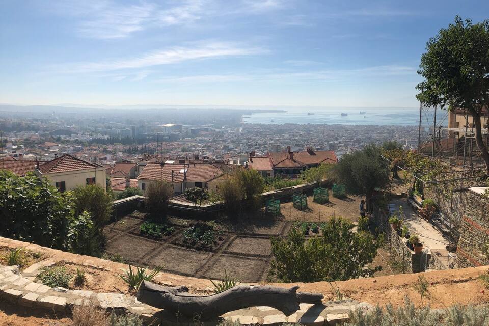 A hillside with terraced gardens, a stone bridge, and a view of a coastal cityscape.