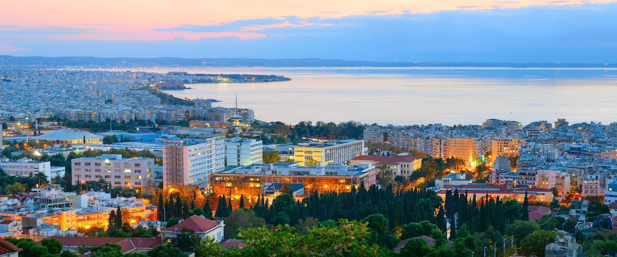 A coastal cityscape at dusk with a prominent waterfront, dense urban buildings, and a historic fortress.