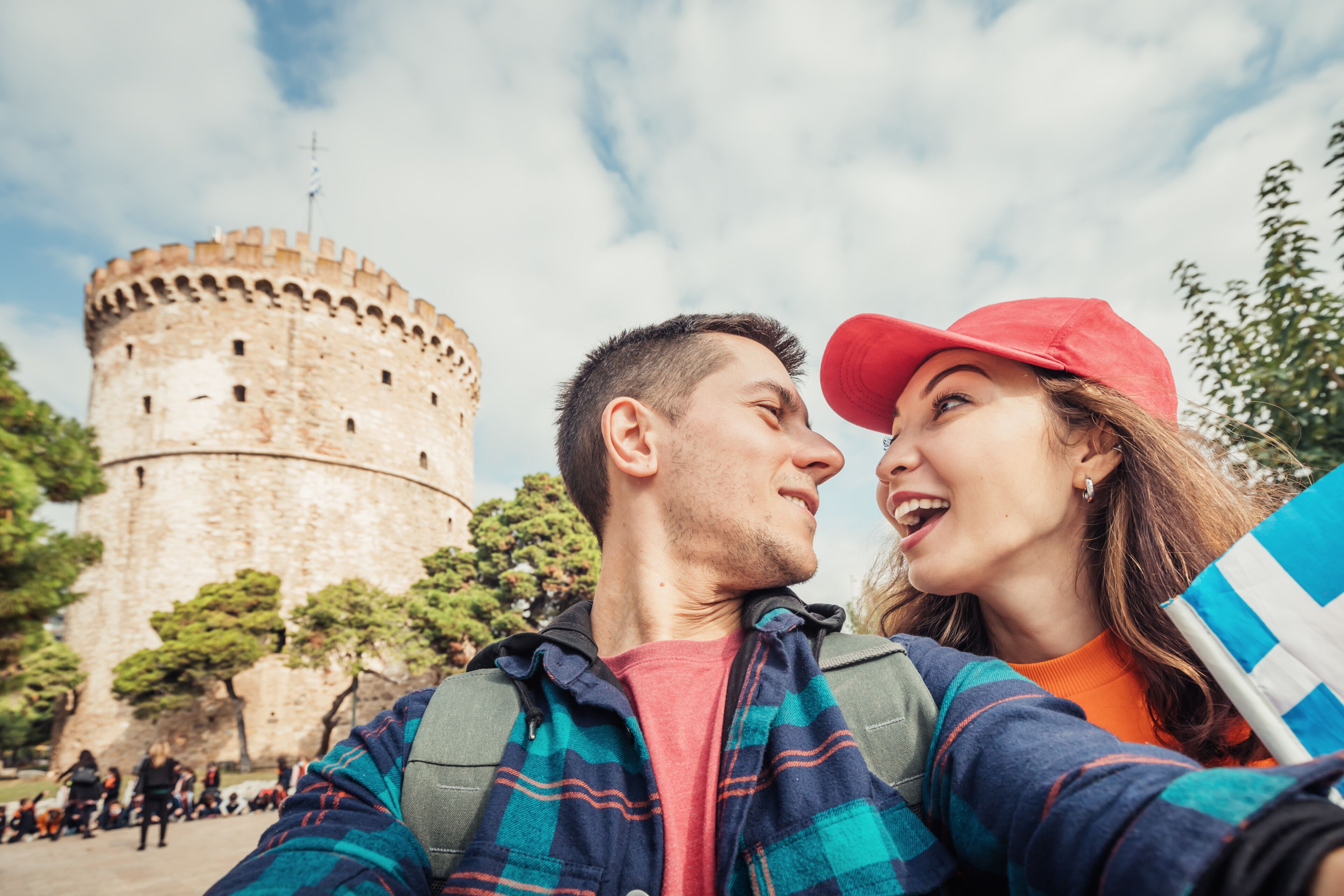 A historic stone tower, a person holding a flag, and a person taking a selfie.