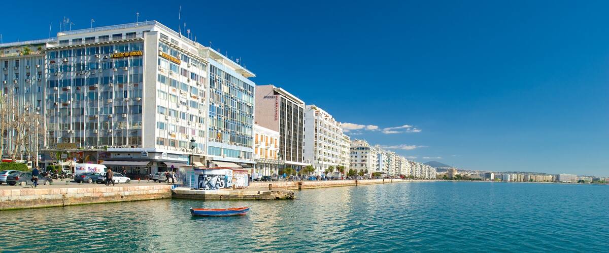 A waterfront cityscape with modern buildings, a calm sea, and a clear blue sky.