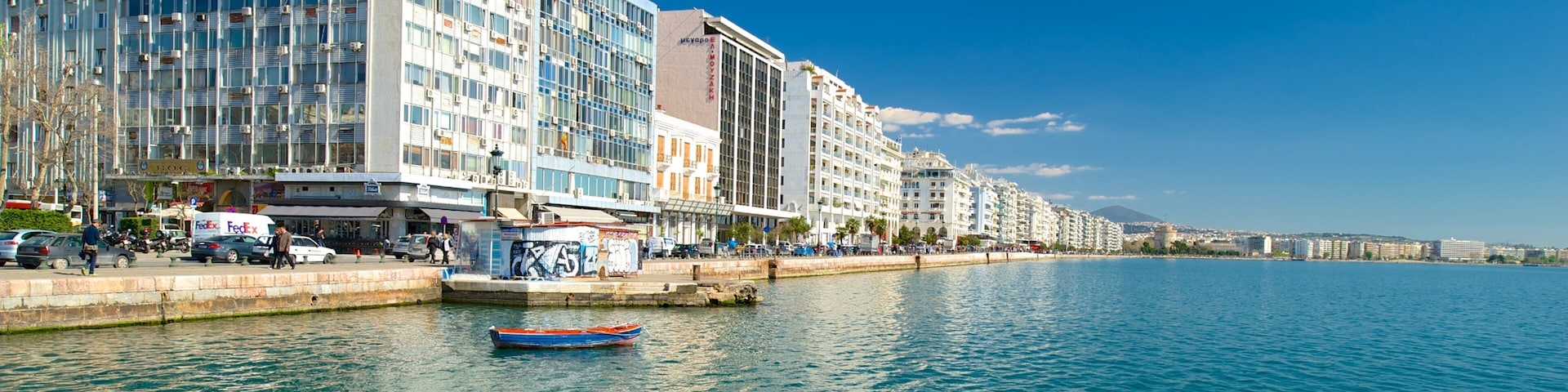 A waterfront cityscape with modern buildings, a calm sea, and a clear blue sky.