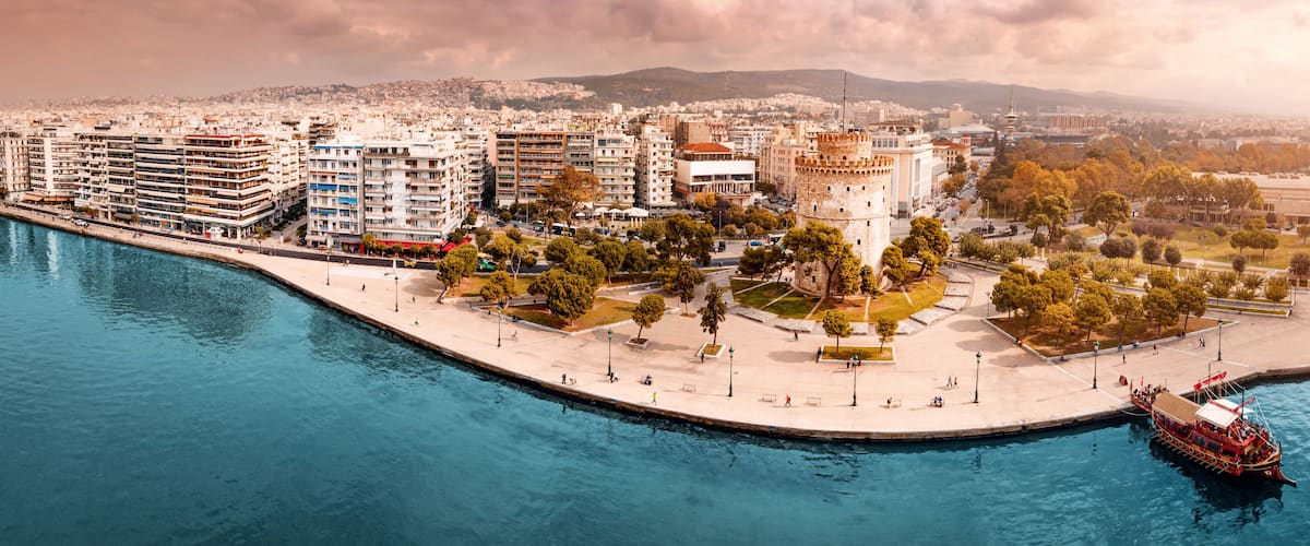 Aerial scenic panorama of the main symbol of Thessaloniki city - the White Tower with boat tour ship at the pier. Concept of travel landmarks in Greece and urban development.