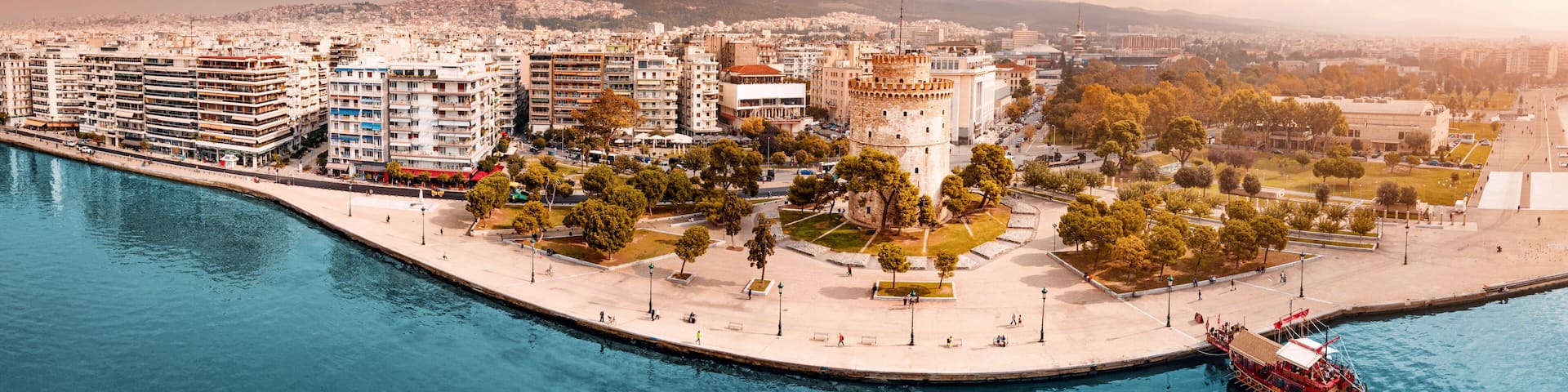 Aerial scenic panorama of the main symbol of Thessaloniki city - the White Tower with boat tour ship at the pier. Concept of travel landmarks in Greece and urban development.