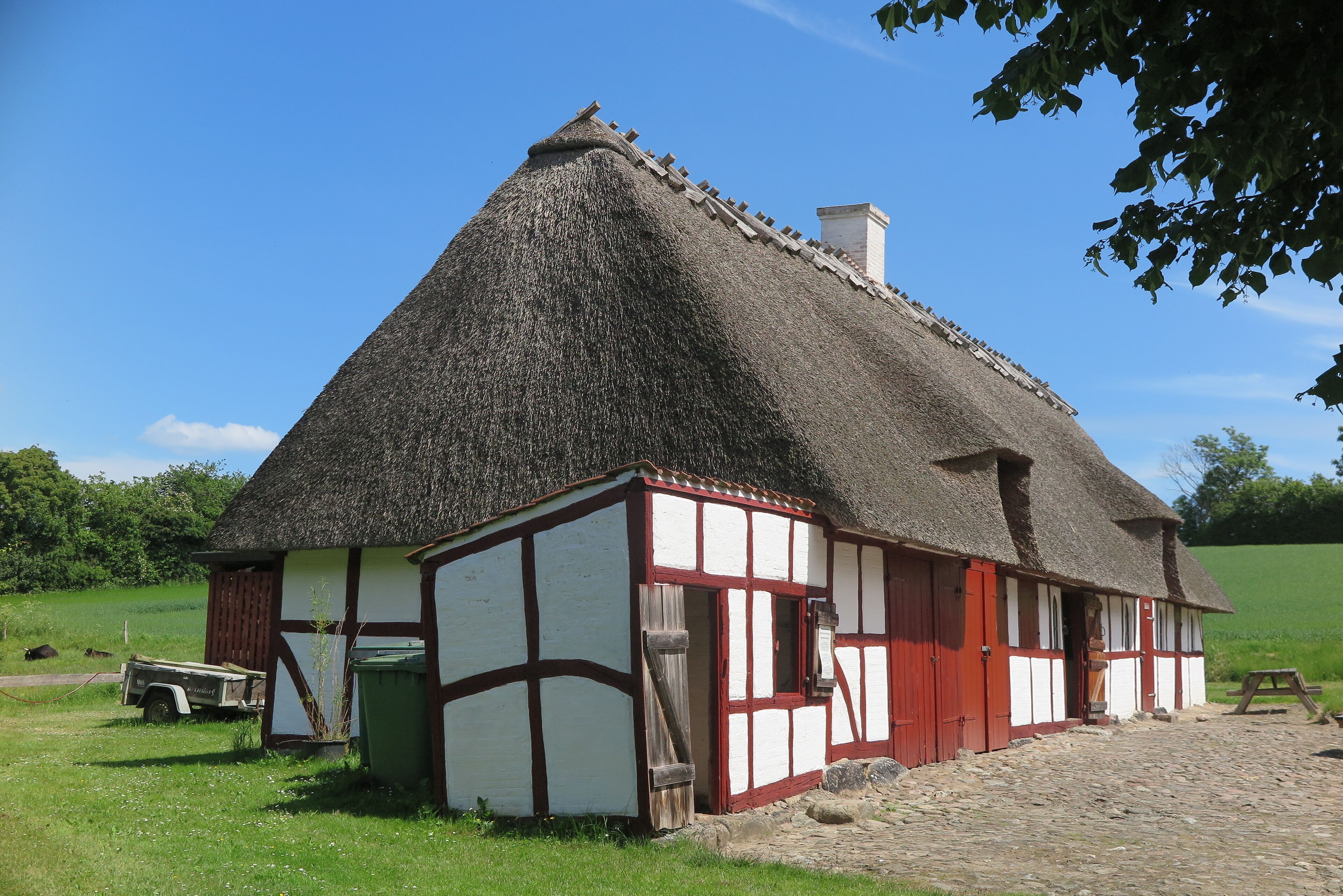 A traditional thatched-roof house with white and red timber framing, set in a rural landscape with a clear blue sky.