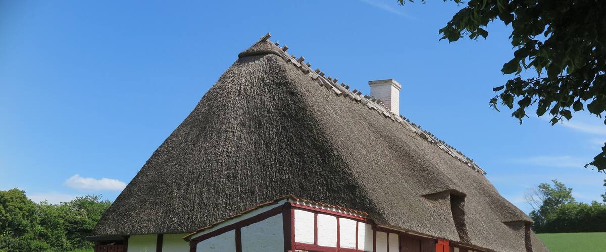 A traditional thatched-roof house with white and red timber framing, set in a rural landscape with a clear blue sky.