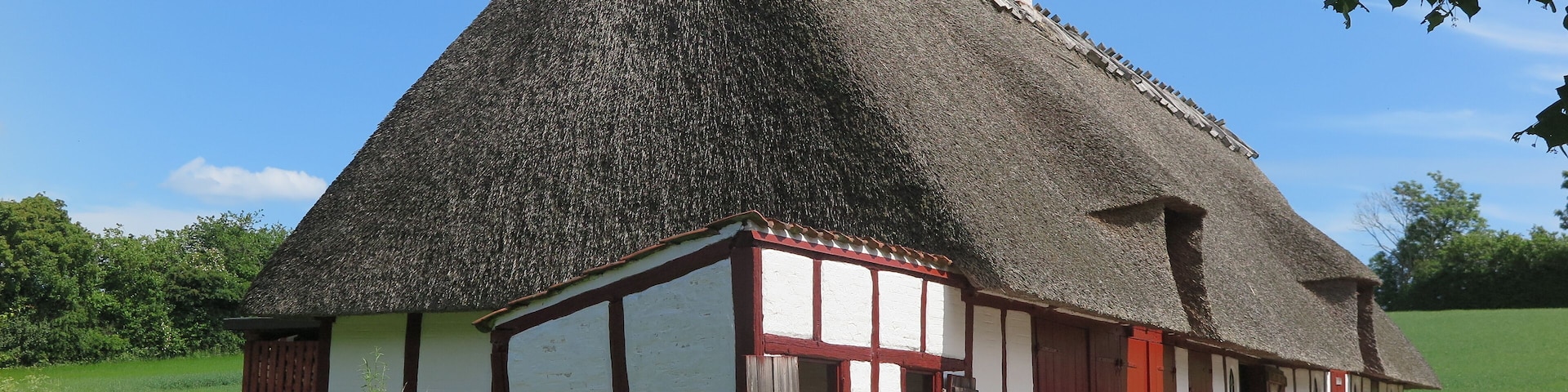 A traditional thatched-roof house with white and red timber framing, set in a rural landscape with a clear blue sky.