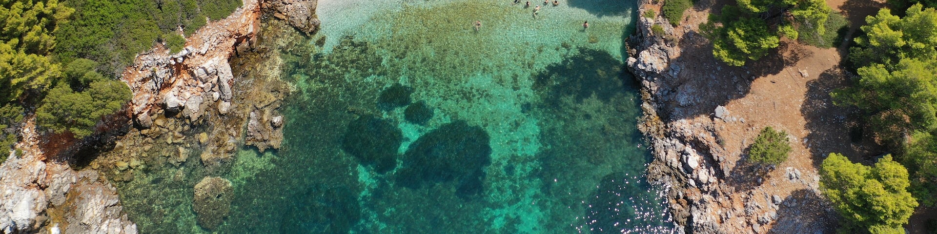 An aerial view of a beach with clear turquoise water, surrounded by dense green trees and rocky terrain.