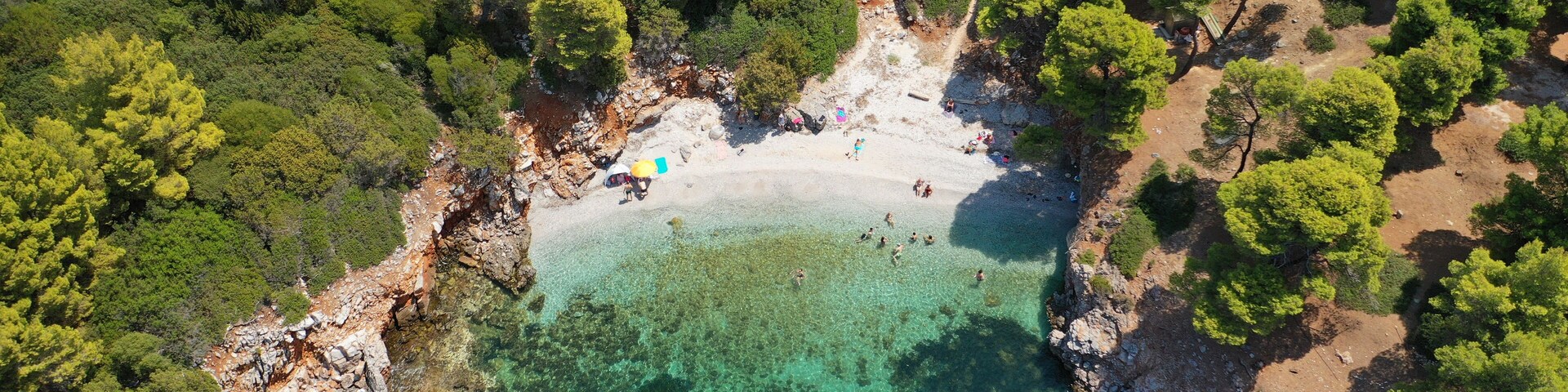 Vista aérea de una playa con aguas turquesas cristalinas, rodeada de árboles verdes y terreno rocoso.