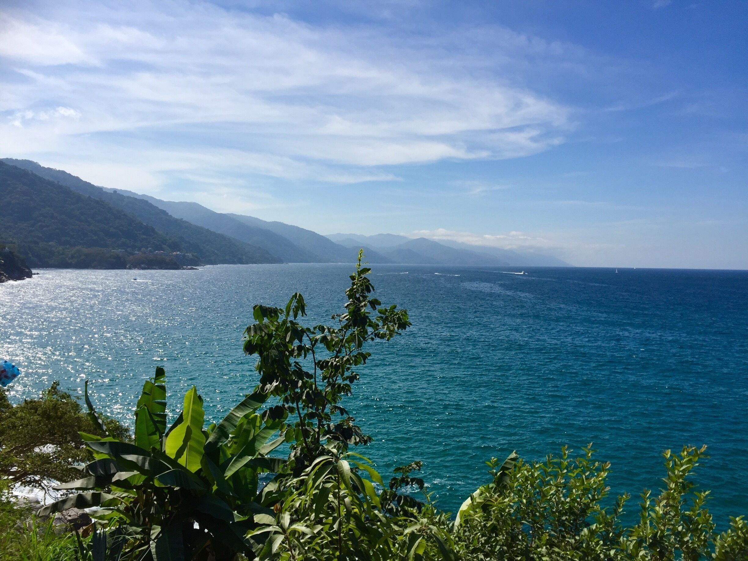 A coastal landscape with a clear blue sea, lush greenery, and distant mountains.