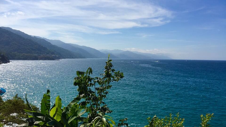 A coastal landscape with a clear blue sea, lush greenery, and distant mountains.