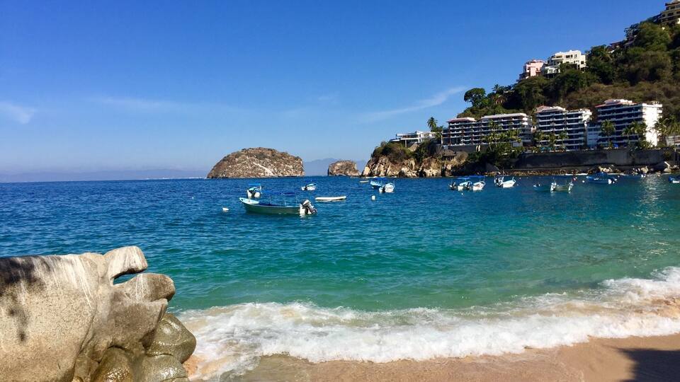 A coastal scene with clear blue waters, a sandy beach, and several boats anchored near rocky outcrops.