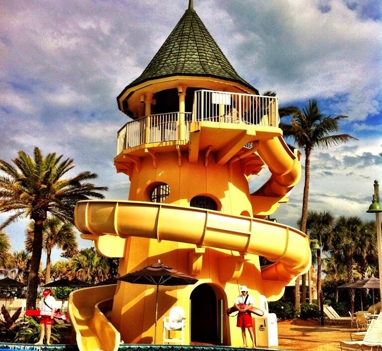 A multi-level water slide structure with a conical roof, surrounded by palm trees and lounge chairs.