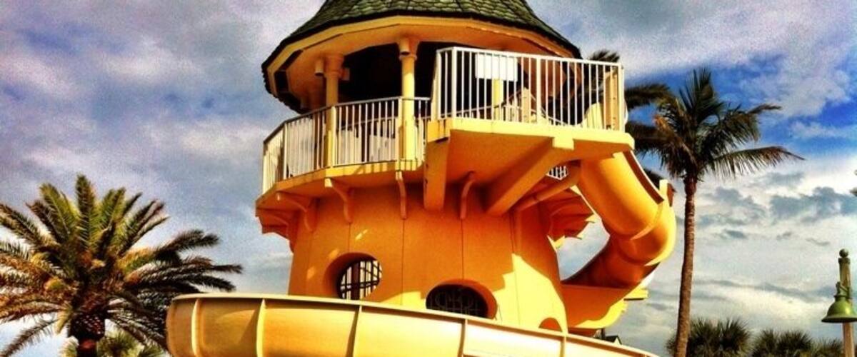 A multi-level water slide structure with a conical roof, surrounded by palm trees and lounge chairs.