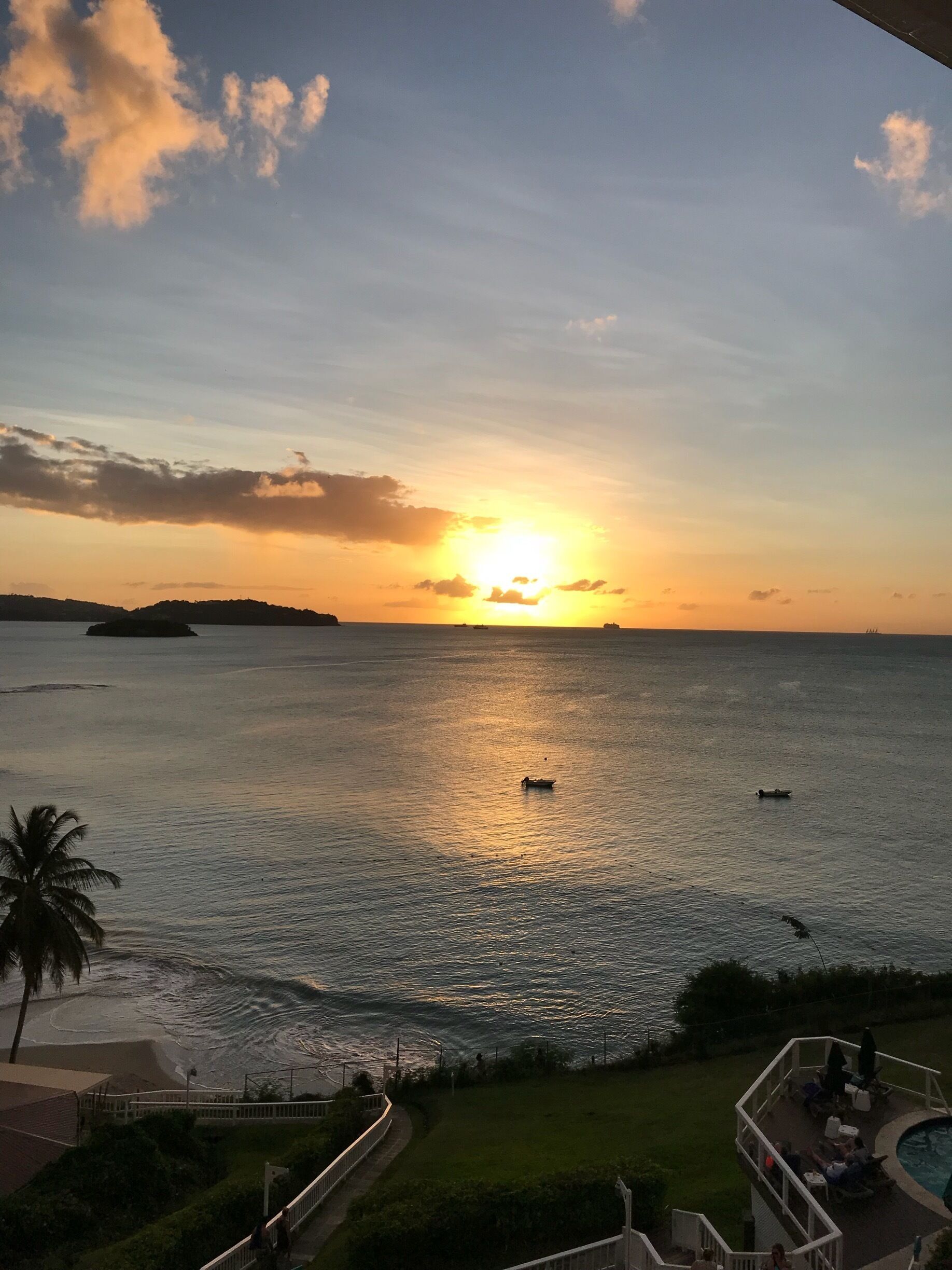 A sunset over a calm sea with boats, a palm tree, and a residential area with a pool.