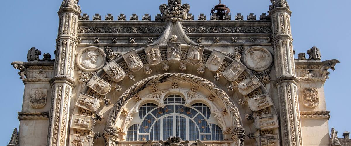 An ornate architectural facade with intricate carvings, a prominent arched window, and a crest at the center.