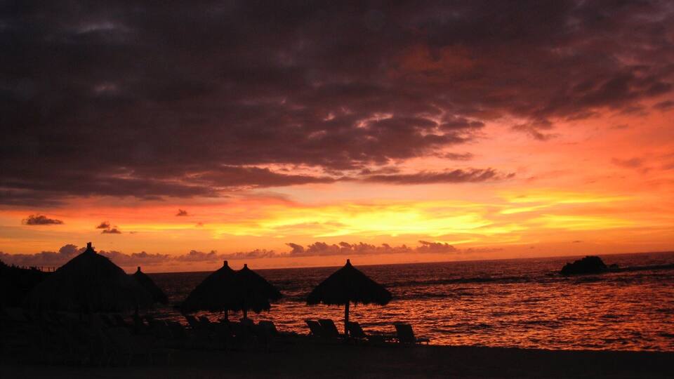 A beach at sunset with thatched huts and lounge chairs.