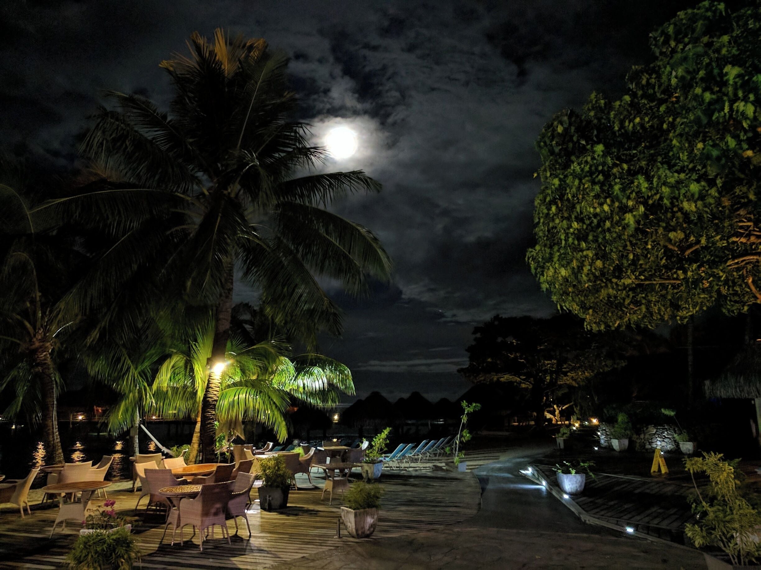 Un espacio al aire libre con mesas y sillas por la noche, con palmeras y una terraza de madera.
