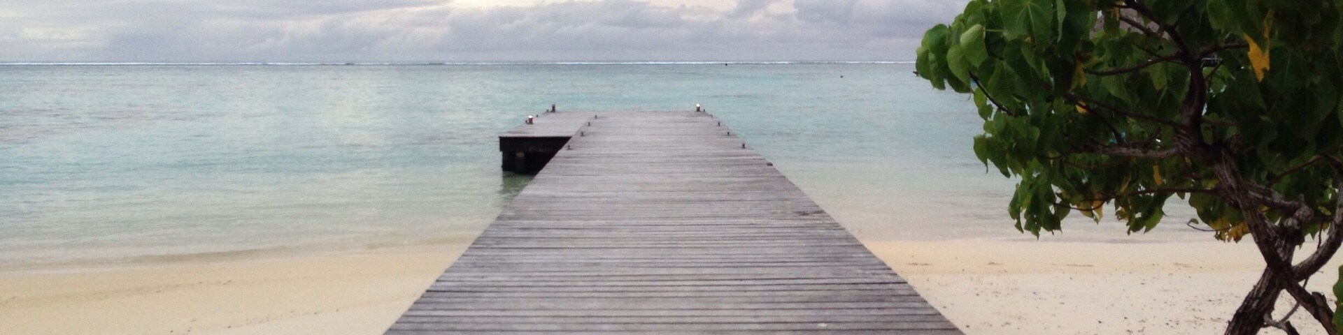 Un muelle de madera se adentra en un mar tranquilo, con una playa arenosa y un cielo nublado.