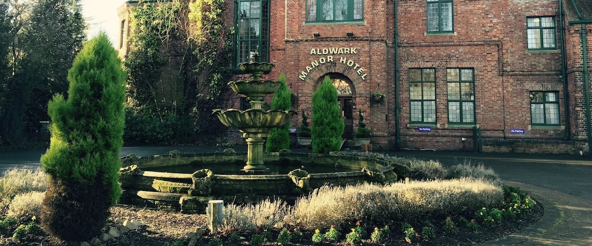 A historic brick building labeled 'Aldwark Manor Hotel' with a fountain and well-maintained garden.