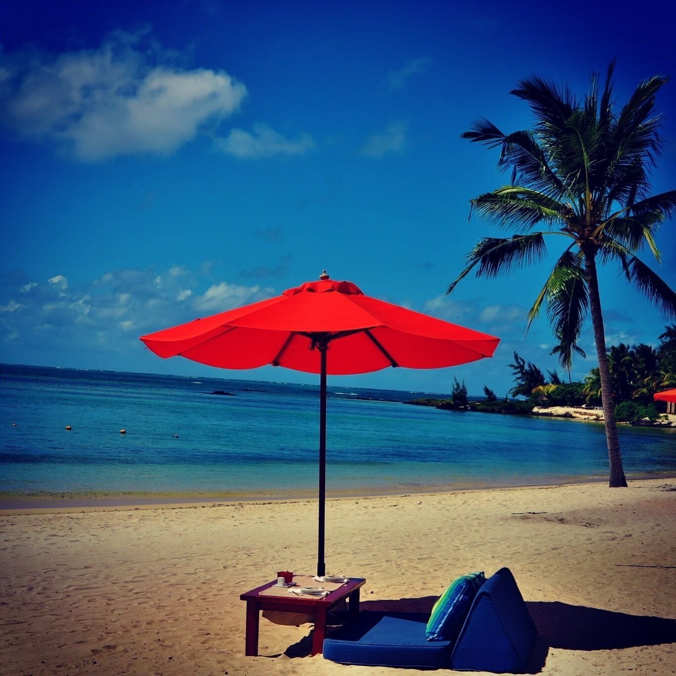 A beach with a red umbrella, a lounge chair, and a palm tree.