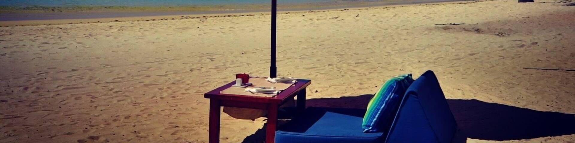 A beach with a red umbrella, a lounge chair, and a palm tree.