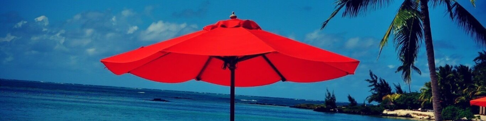 Uma praia com um guarda-sol vermelho, uma espreguiçadeira e uma palmeira.