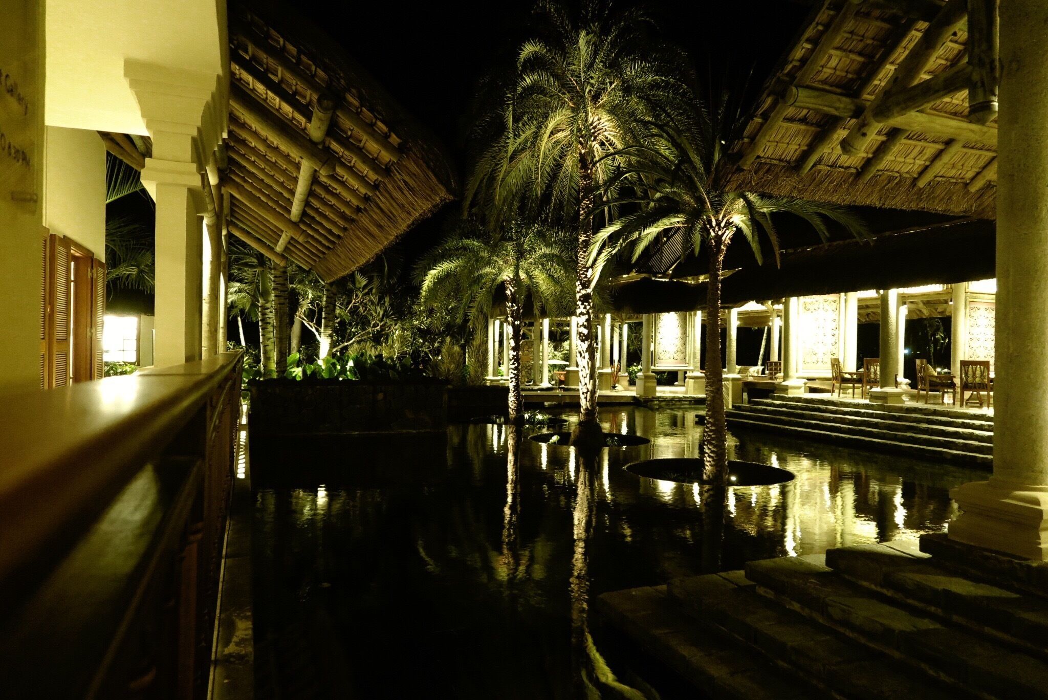 A nighttime resort with a pool, palm trees, and thatched-roof buildings.