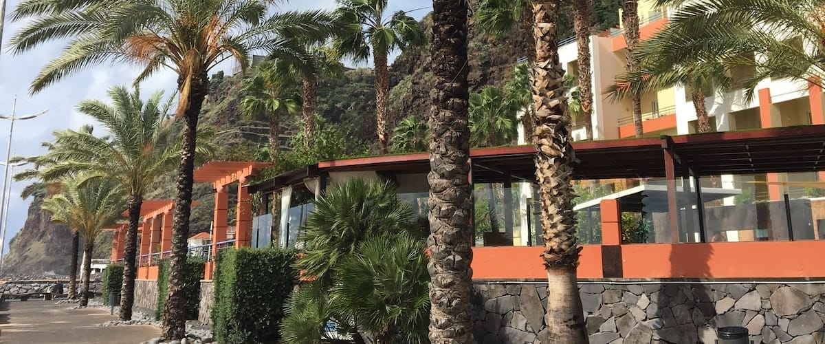 A coastal area with palm trees, a paved walkway, and buildings with orange and white facades.