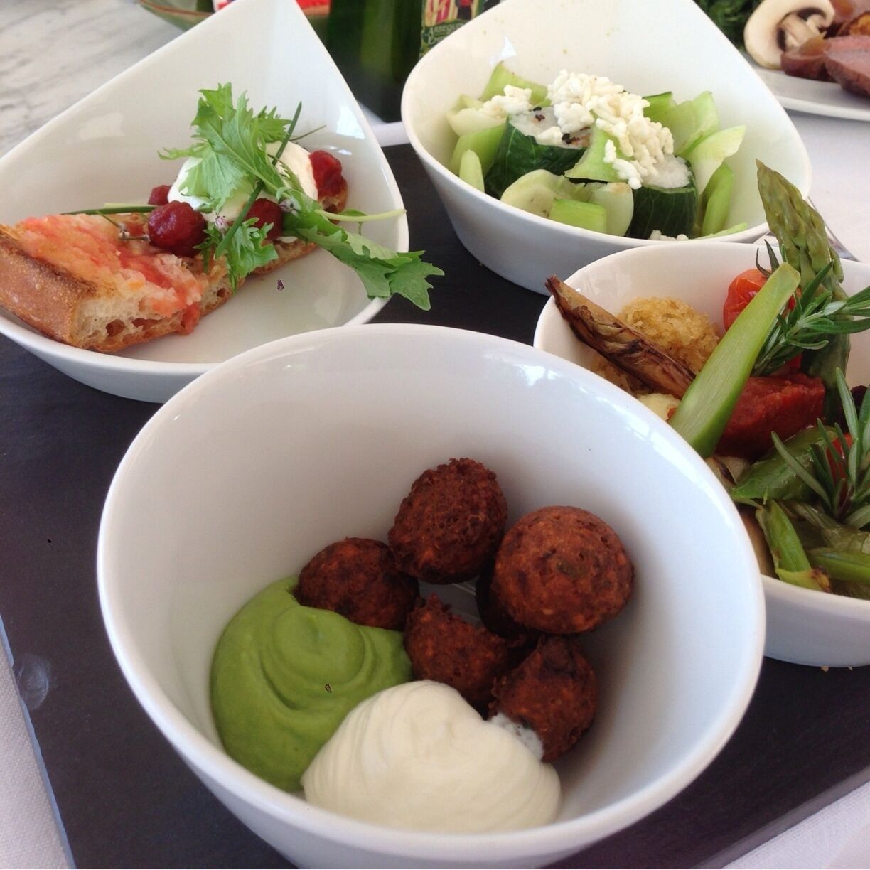 A variety of dishes including a bowl of falafel, avocado, and sauce; a bowl of salad with feta cheese; a bowl of sliced cucumbers and tomatoes; and a plate of meat with greens.