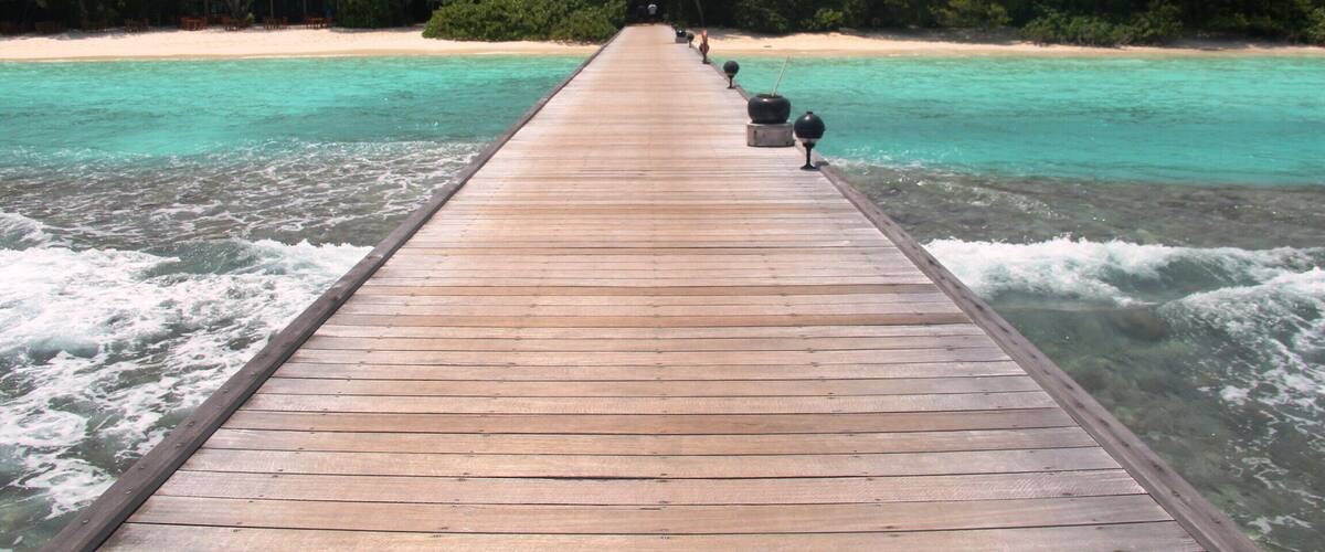 Un muelle de madera que se adentra en aguas turquesas, con una playa y abundante vegetación en el fondo.