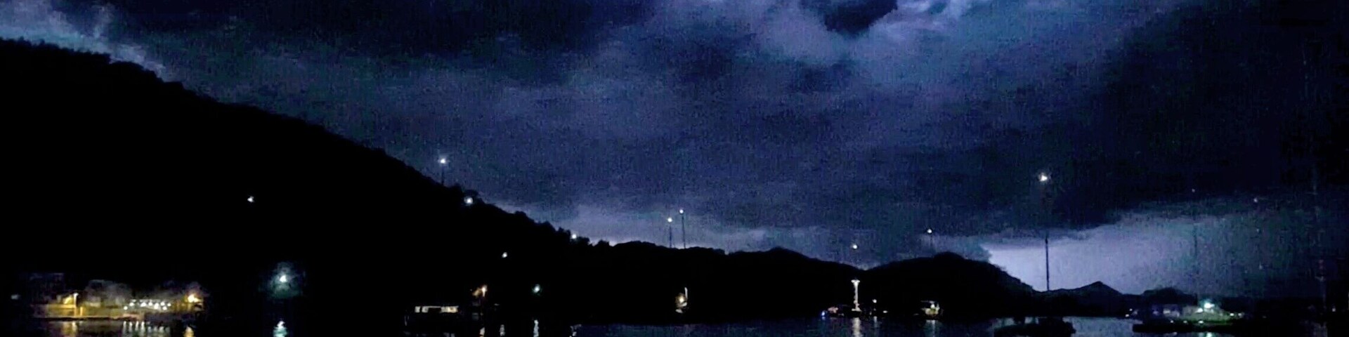 Escena nocturna junto al lago, con barcos y luces distantes en una colina.