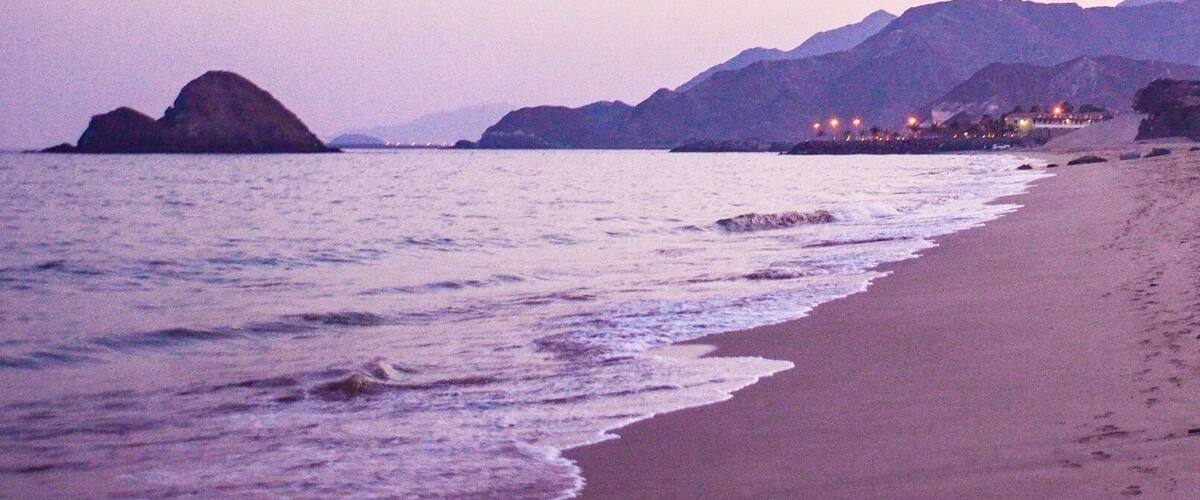 A beach with waves, a large rock formation, and distant mountains.