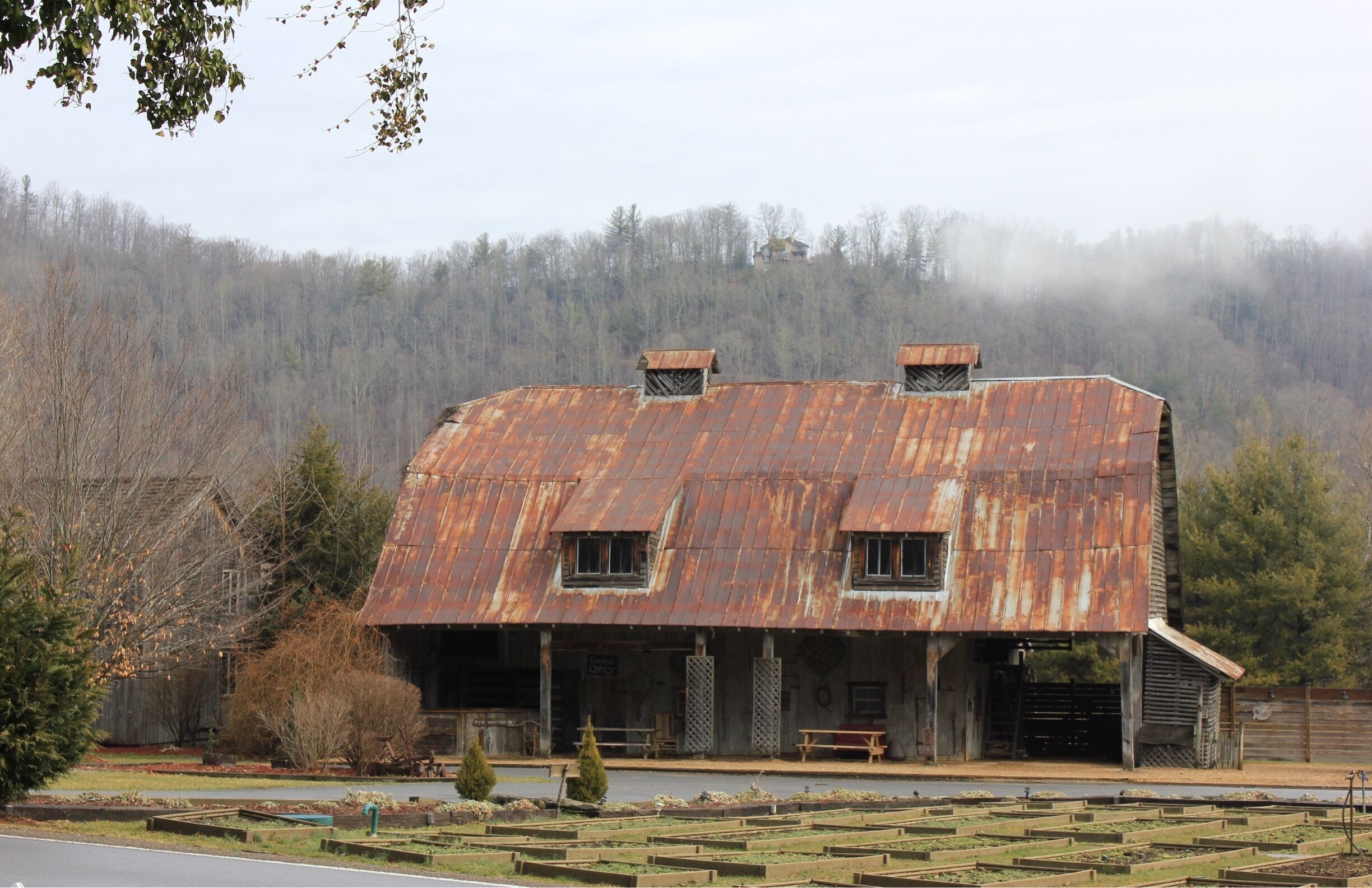 A rustic barn with a rusted metal roof, surrounded by a vegetable garden and a forested hillside in the background.