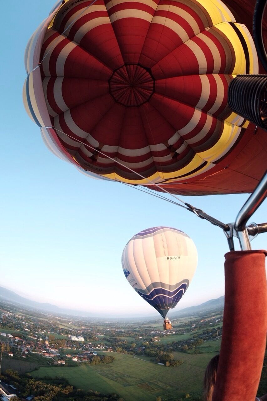 Two hot air balloons, one with red and white stripes and another with blue and white, flying over a rural landscape with fields and houses.