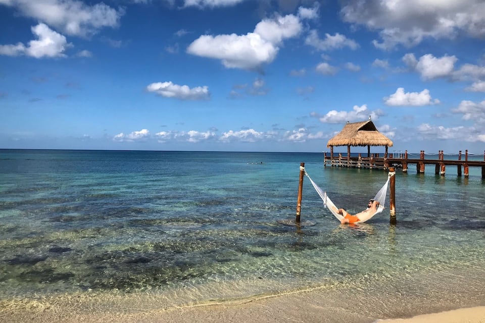 A beach with clear turquoise water, a hammock, a thatched-roof hut, and a wooden pier.