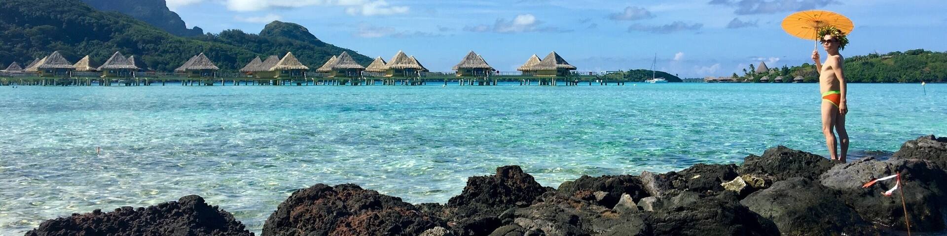 A person standing on rocks by a turquoise sea with overwater bungalows and a mountain in the background.