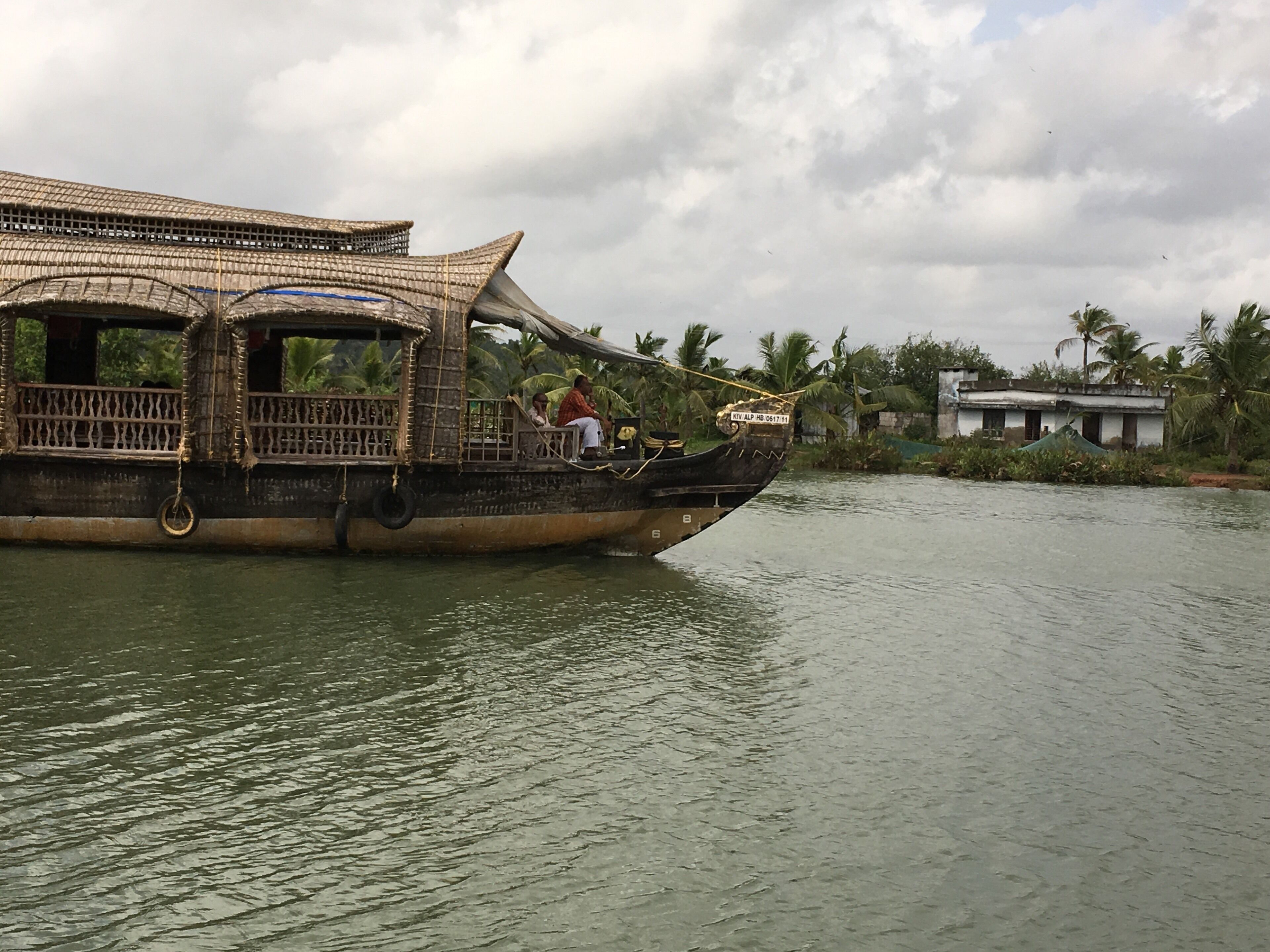 A traditional wooden boat with a thatched roof, passengers, and a house in the background.