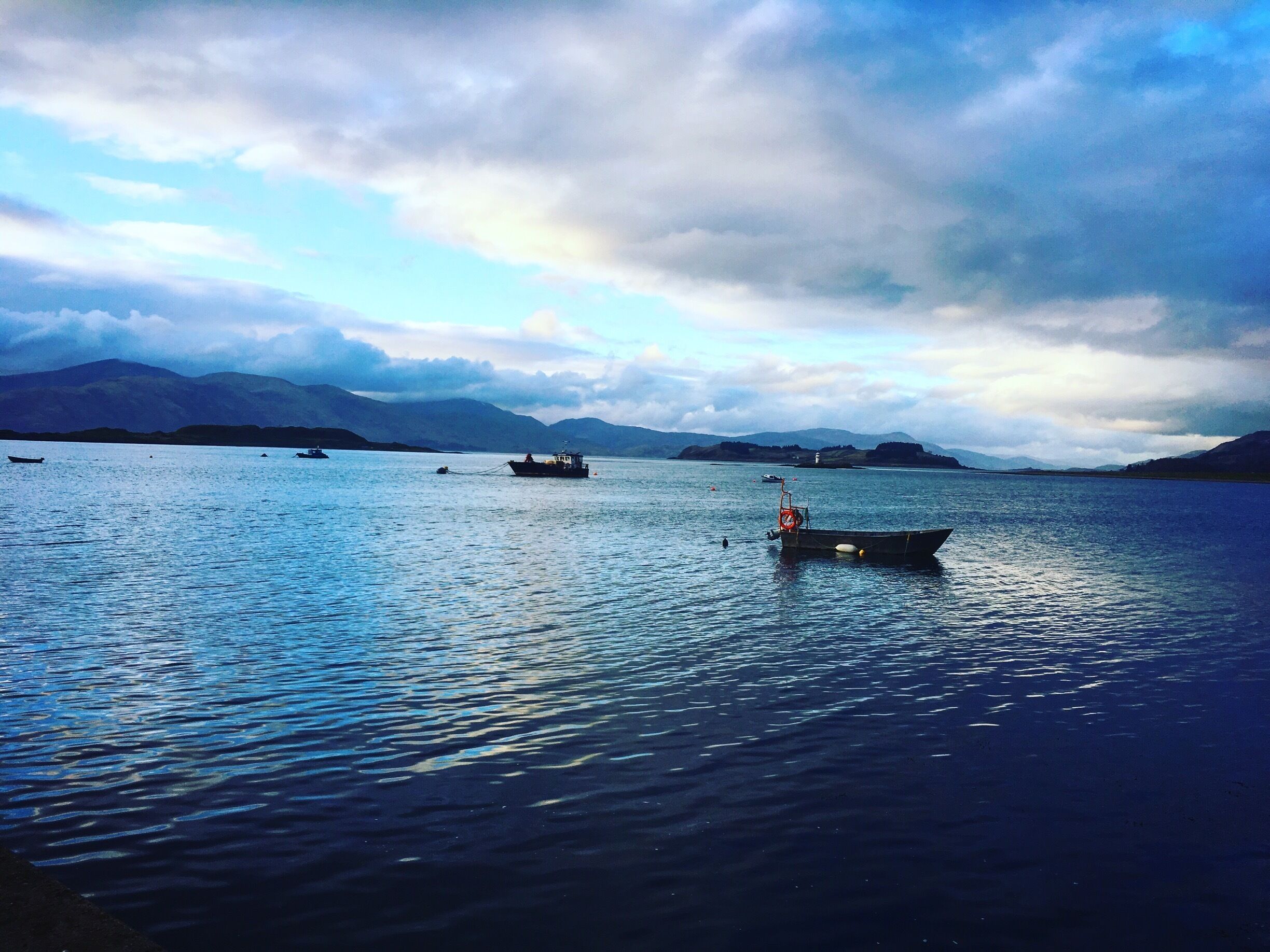 A calm lake with several boats, mountains in the background, and a cloudy sky.