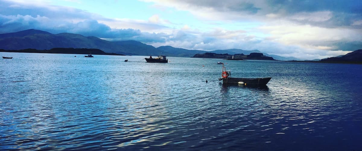 A calm lake with several boats, mountains in the background, and a cloudy sky.