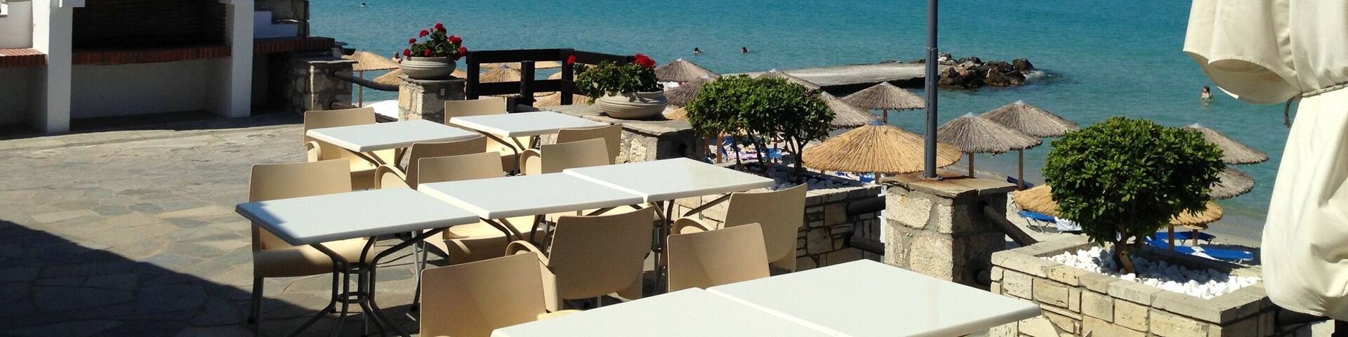 Un patio frente al mar con mesas y sillas, zona de barbacoa y sombrillas con vistas al océano.