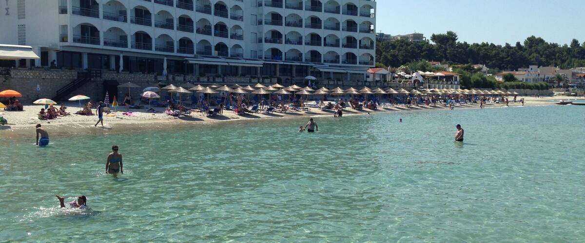 A beach with clear water, umbrellas, and a large hotel building.