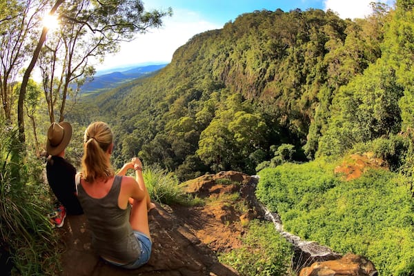 Two people sitting on a rocky ledge overlooking a lush valley with a waterfall.